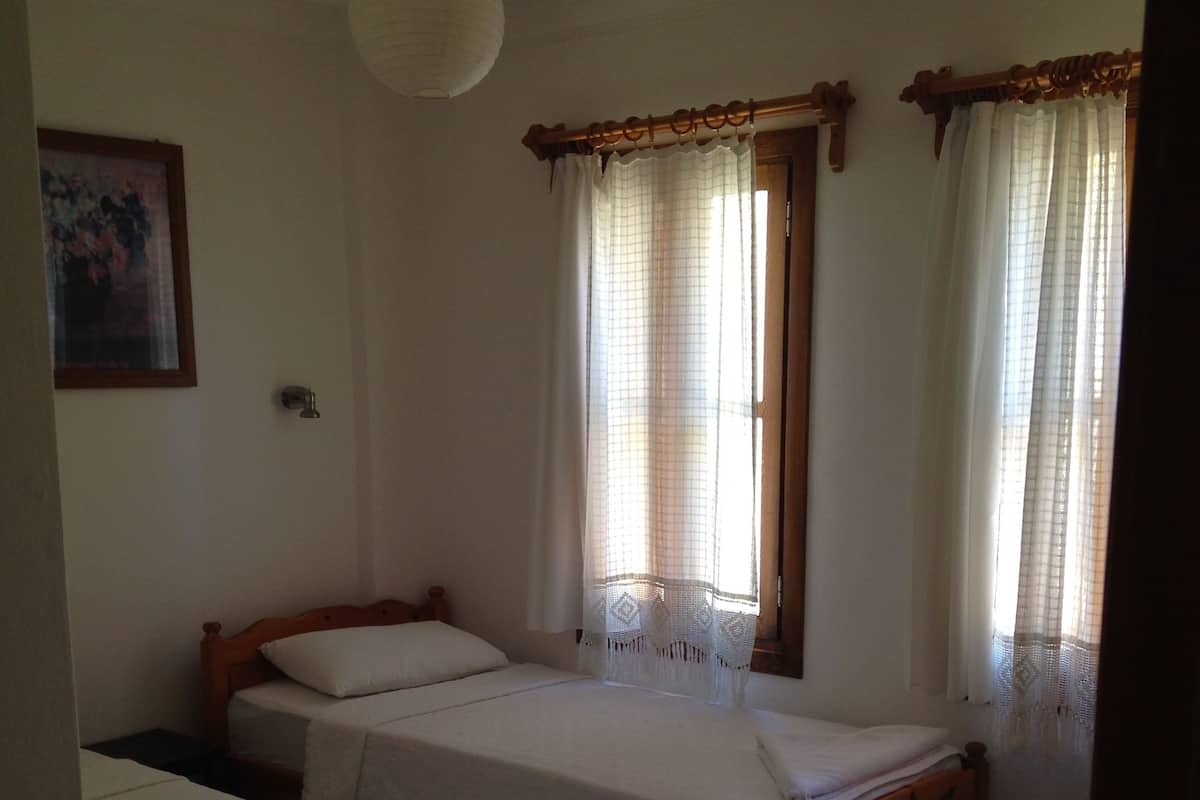 Een slaapkamer met twee eenpersoonsbedden, een houten hoofdbord, een kamerkleed met patroon en een raam met dunne gordijnen.