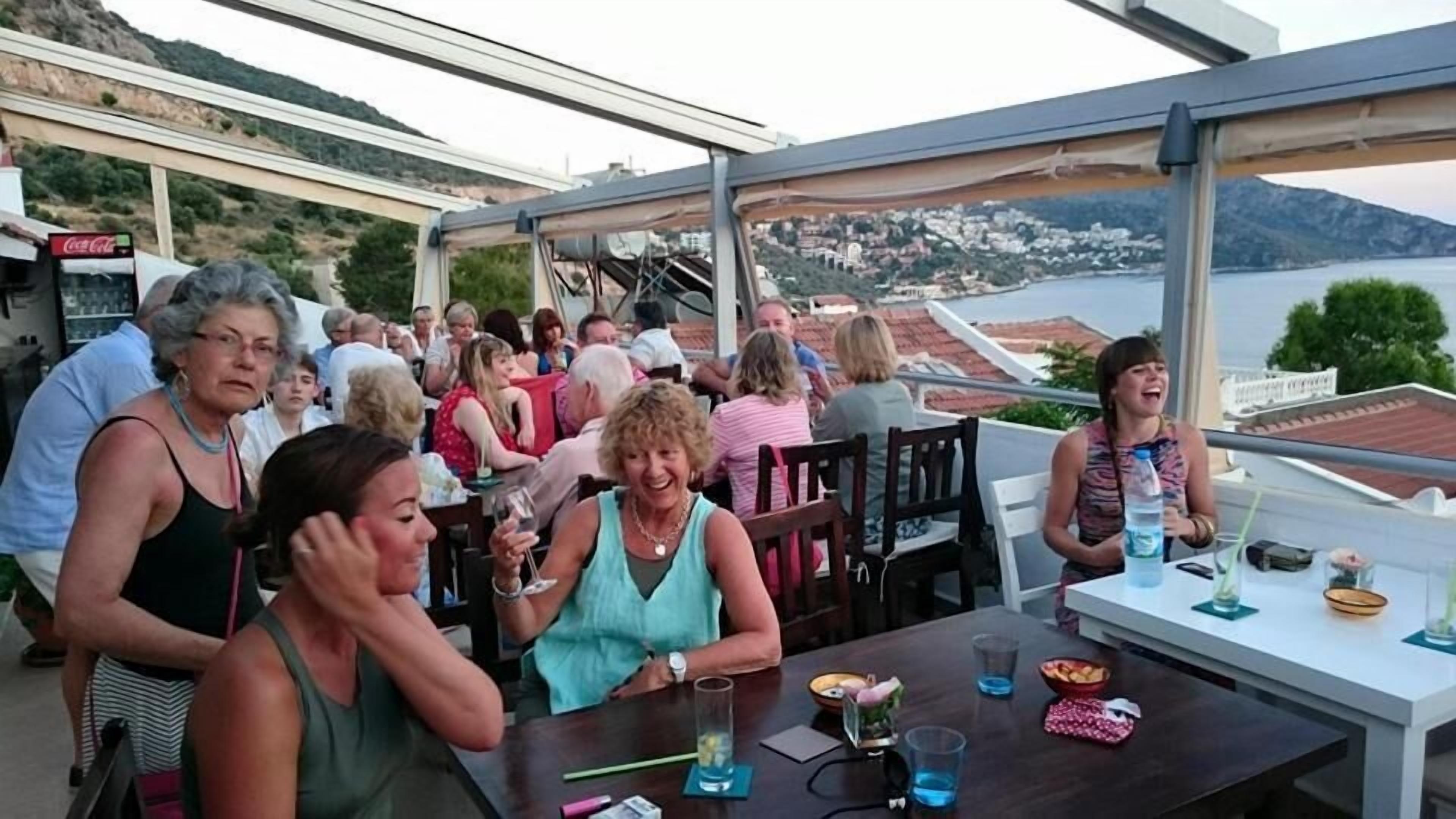 Um grupo de pessoas sentadas numa mesa ao ar livre, com vista para uma vila costeira e colinas ao fundo.