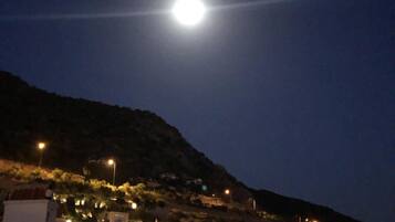 Cena noturna com uma lua brilhante no céu, um morro com edifícios e postes de luz.