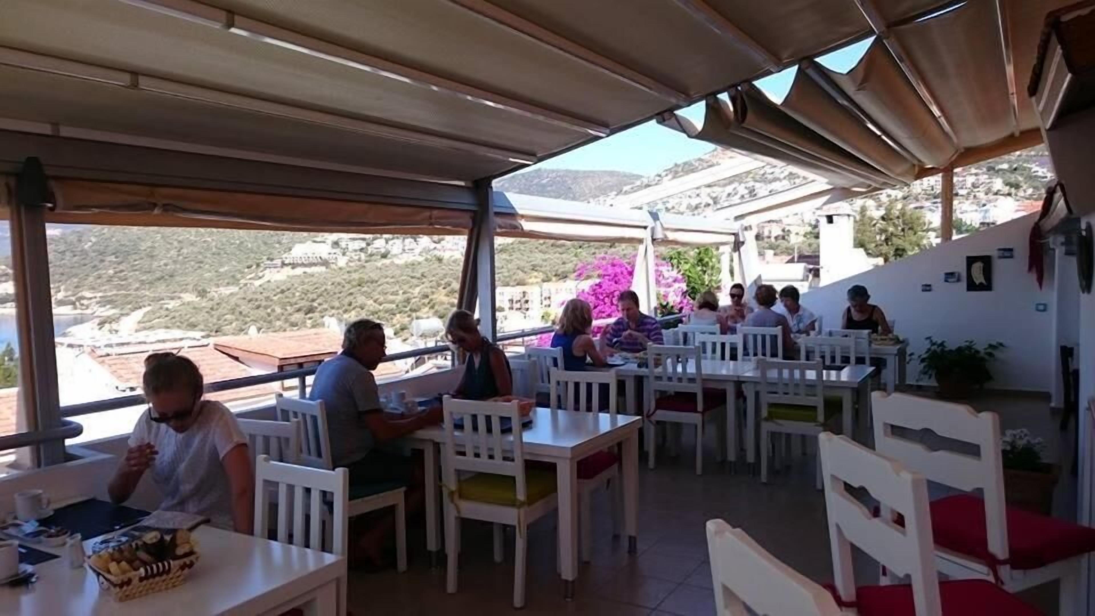 Un café al aire libre con mesas y sillas blancas, vista a un paisaje montañoso y un área cubierta con techo.