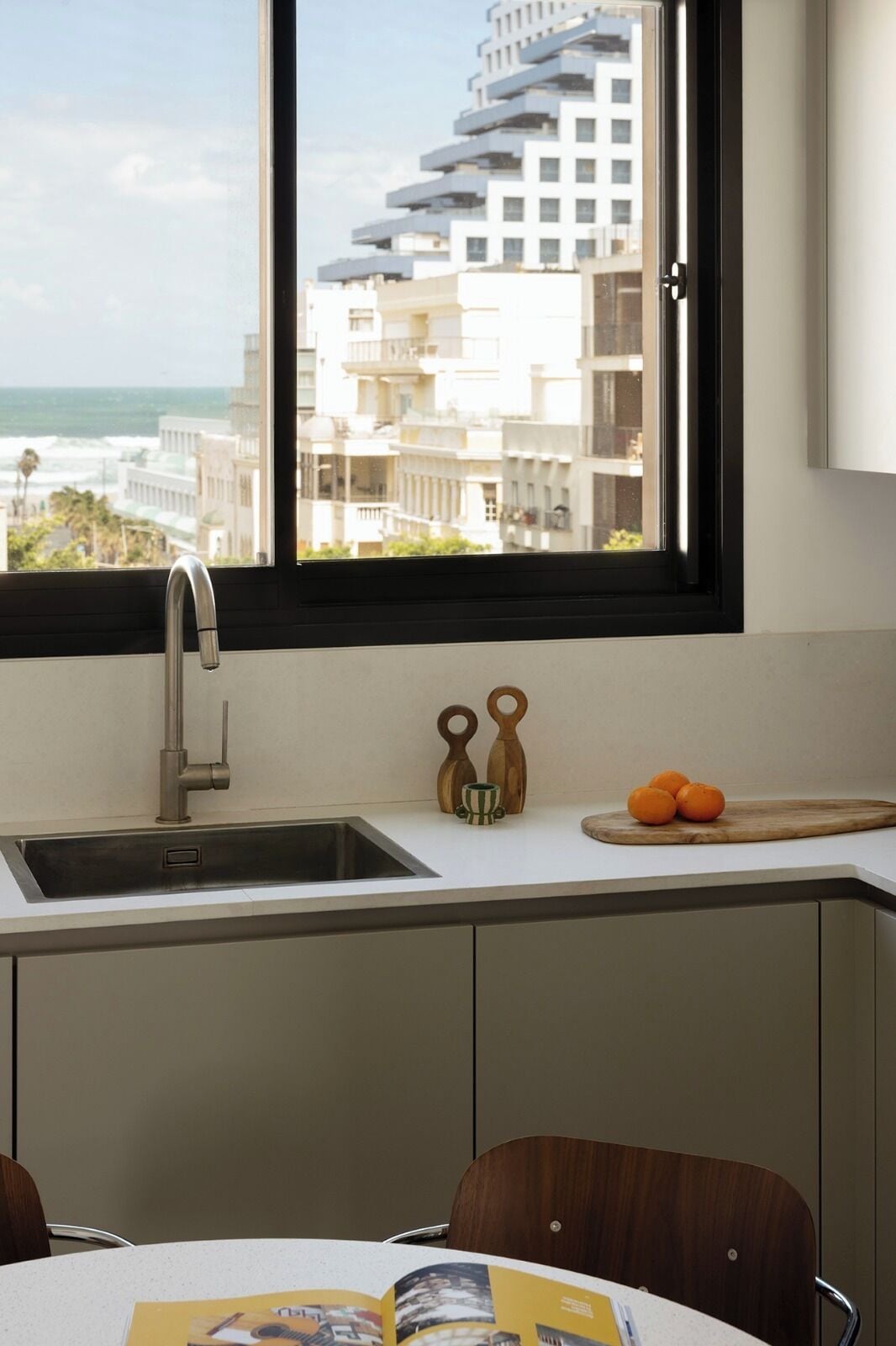 A modern kitchen with a large window, a stainless steel sink, and a wooden cutting board with oranges.