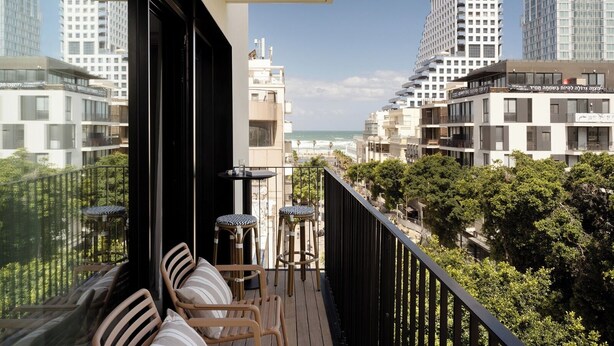 A balcony with wicker chairs and a small table, overlooking a cityscape with modern buildings and a view of the sea.