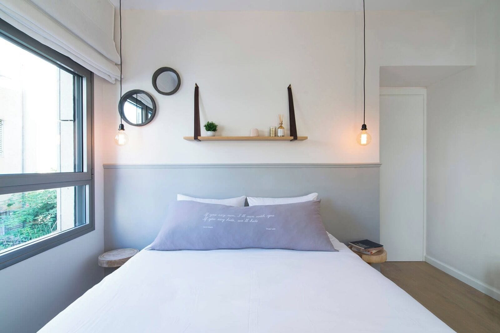 A bedroom with a bed, a window, a bedside table, a shelf with decorative items, and wall-mounted mirrors.
