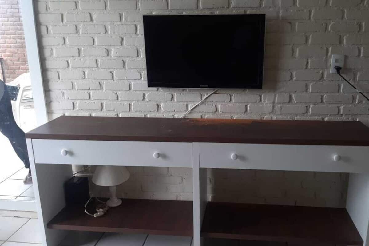 A white brick wall with a flat-screen TV mounted above a wooden desk with two drawers and a shelf.