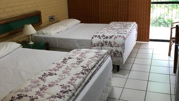 A hotel room with two beds, a wooden headboard, a green nightstand, and a view of greenery outside. - Flat Angra dos Reis Portogalo (Angra dos Reis)