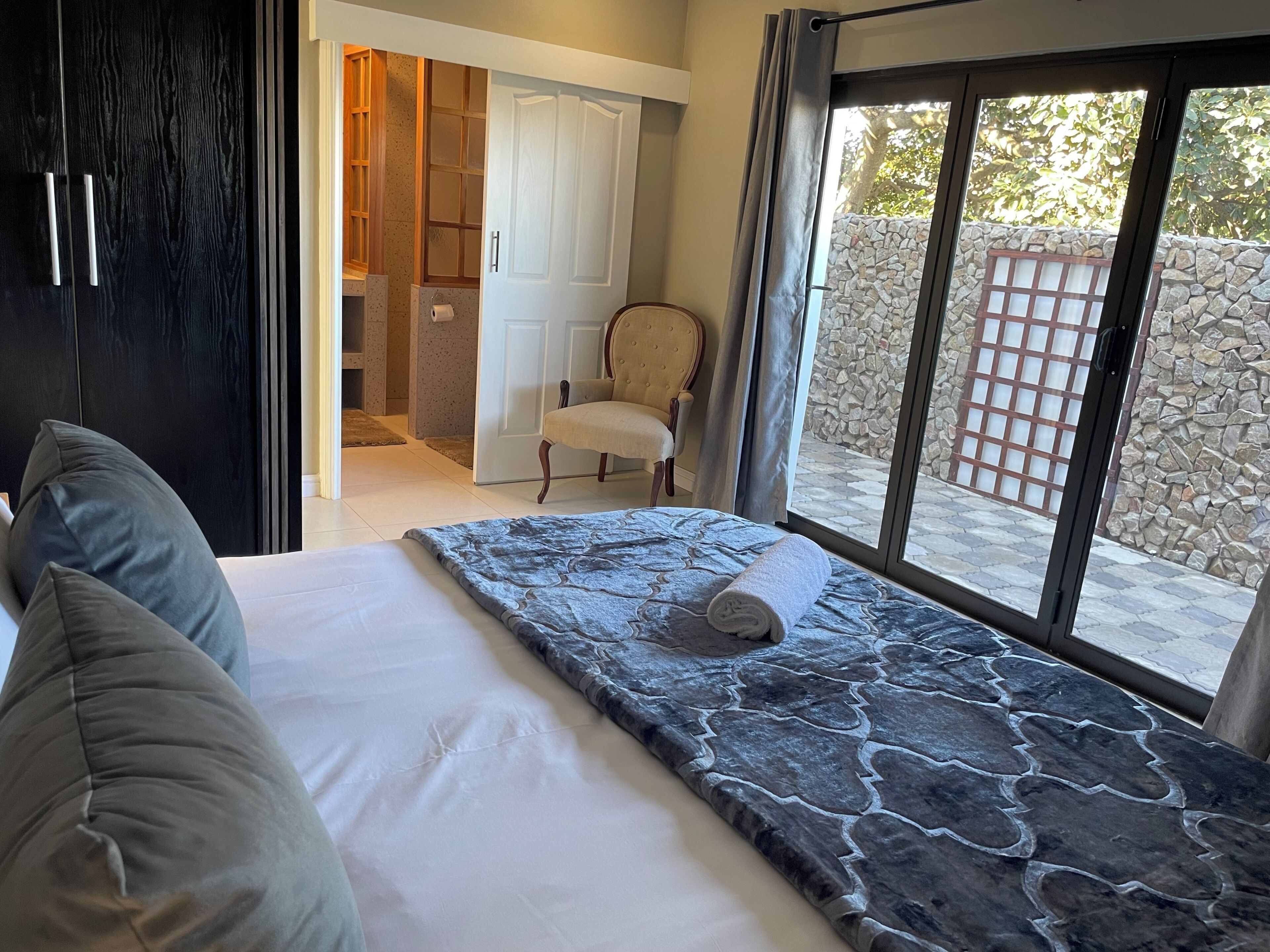 A bedroom with a bed, pillows, a chair, a wardrobe, and a view of the outside.