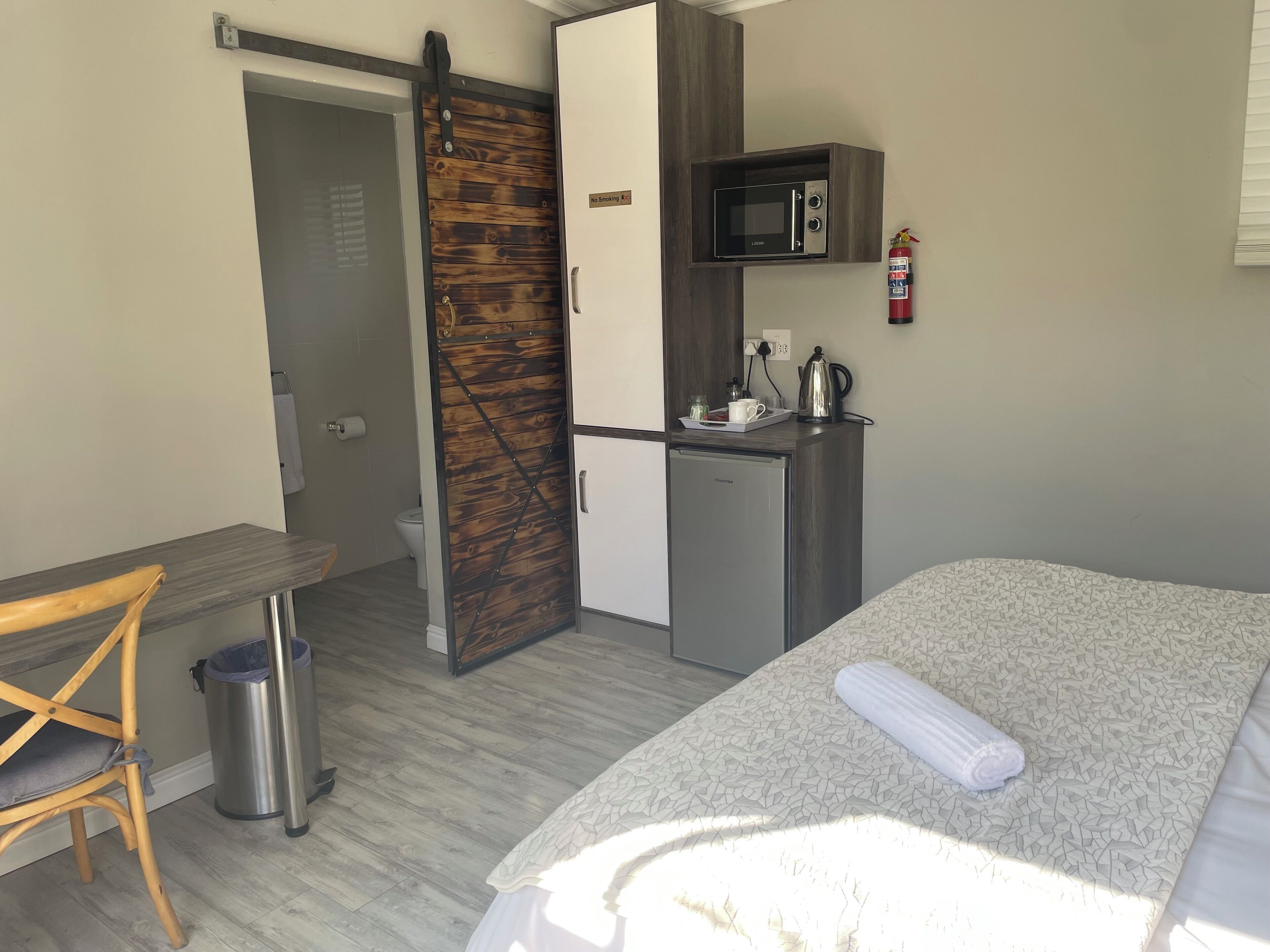 A hotel room with a bed, a desk, a kitchenette with a microwave, a trash bin, and a sliding door.