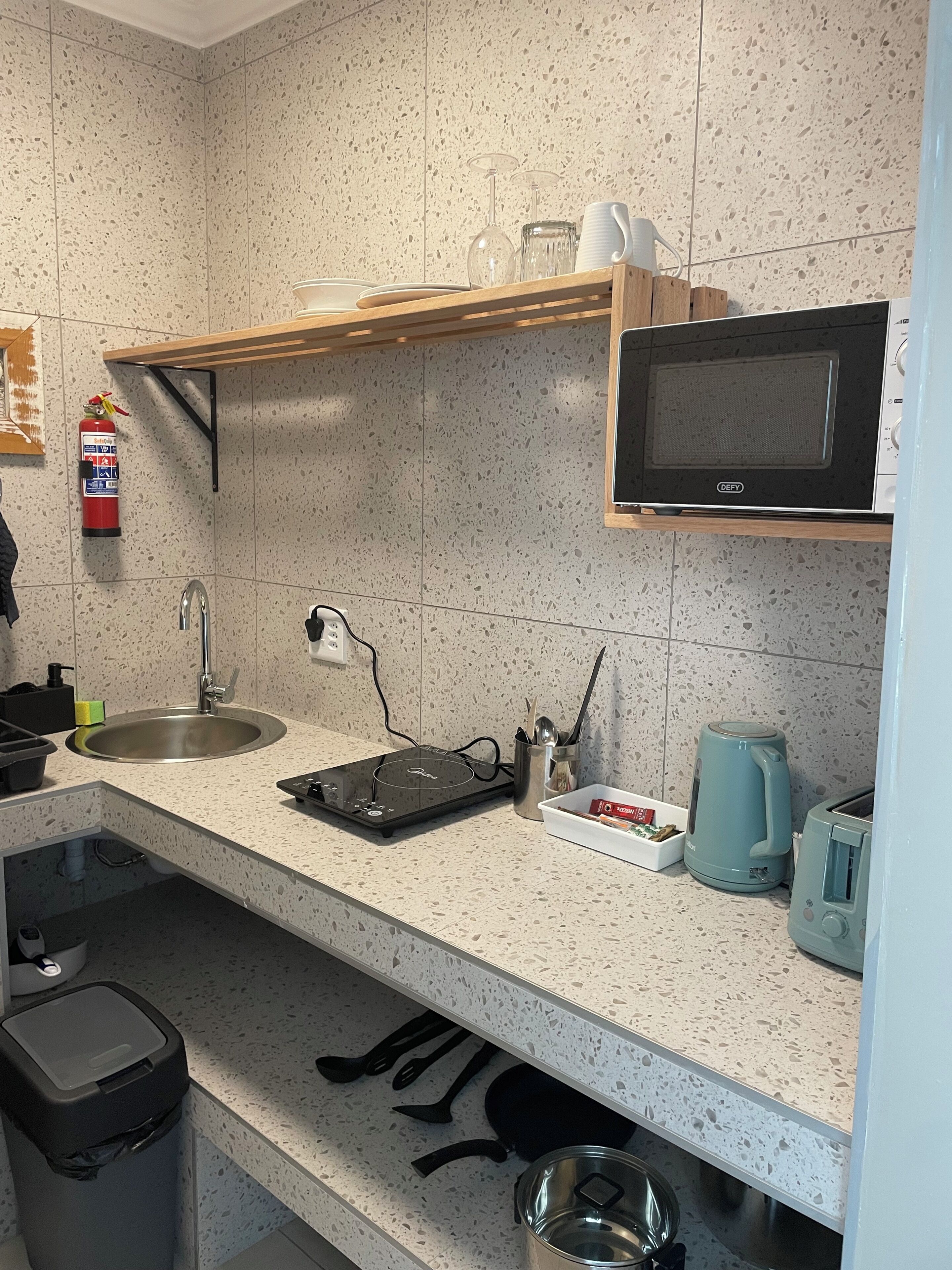 A kitchenette with a microwave, kettle, toaster, sink, and a small shelf with cups and a fire extinguisher.