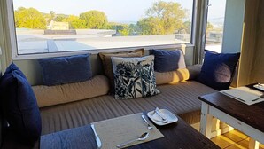 Une pièce dotée d’une grande fenêtre, d’un canapé avec des coussins, d’une petite table avec un set de table et des couverts, et offrant une vue sur un immeuble et des arbres à l’extérieur.