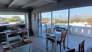 Un espace repas avec des tables et des chaises, de grandes fenêtres offrant une vue sur la ville et un balcon doté d’une balustrade.