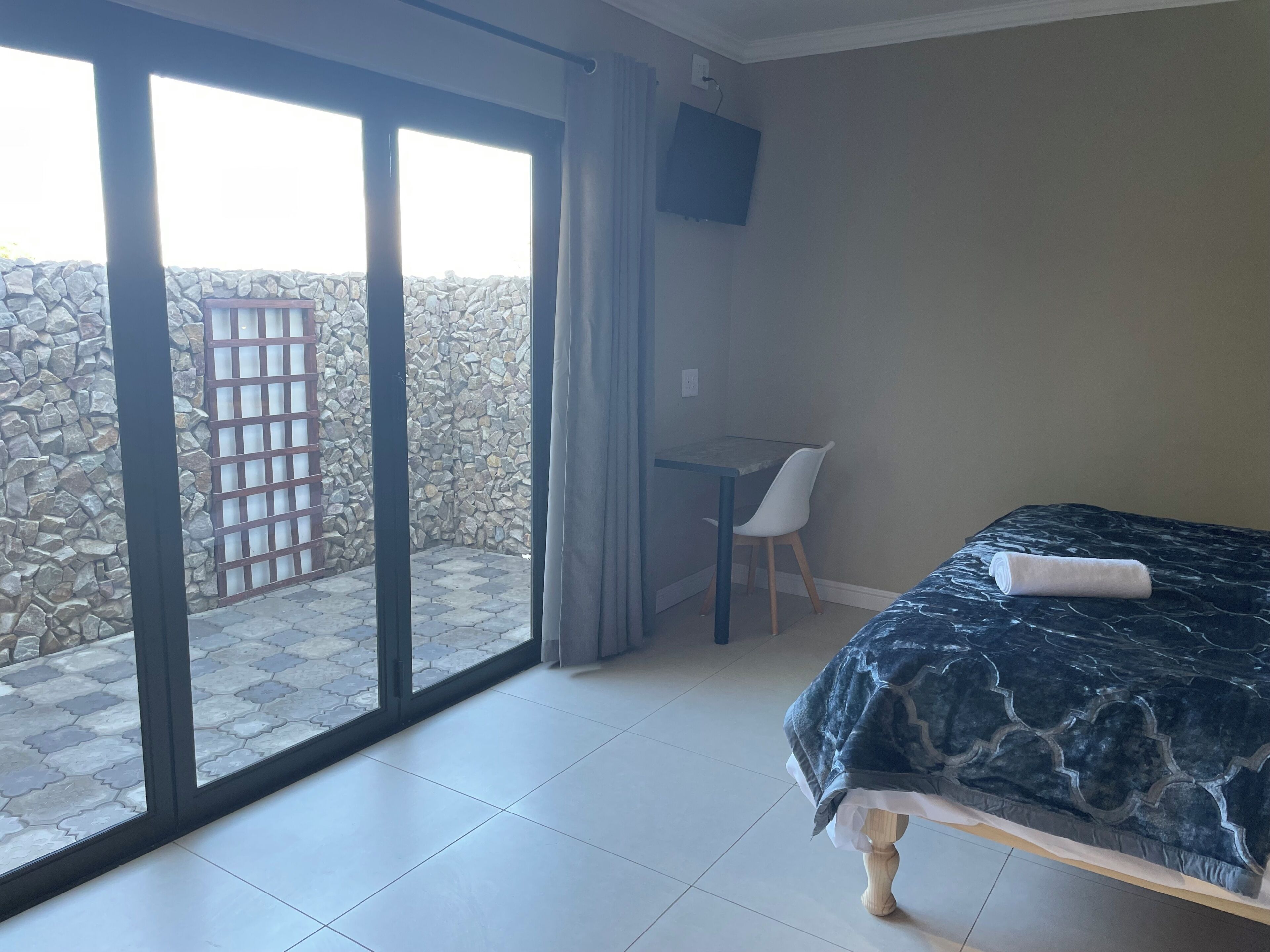 A room with a bed, a desk, a chair, a TV, and a view of a stone wall and a gate.