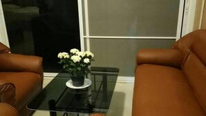 A living room with two brown leather sofas, a glass coffee table with a flower vase, and a large window with blinds. - Hill Tribe Art House (Chiang Rai)
