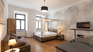 A modern bedroom with a four-poster bed, a wooden floor, a sofa, a small table with a lamp, and a TV mounted on the wall.