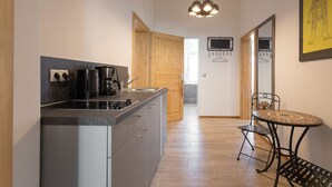 A modern kitchen with a built-in stove, a coffee maker, and a sink. There is a wooden door leading to another room, a small table with a chair, and a hanging TV.