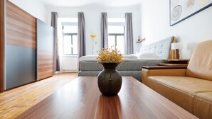A modern hotel room with a wooden table, a vase with flowers, a sofa, a bed, and a window with curtains.