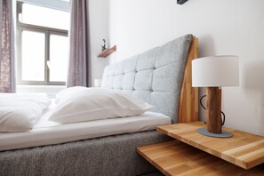 Un lit bien fait, avec du linge de lit blanc et des oreillers, une table de chevet en bois avec une lampe, et une fenêtre avec des rideaux.