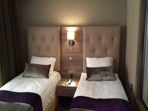 Una camera d'albergo con due letti singoli, ciascuno con cuscino e coperta. Tra i letti si trova un comodino con una lampada.