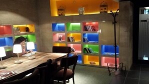 A dining area with a wooden table, chairs, and a person seated. There are colorful shelves with books and decorative items. - Reda Palas Karanfil (Ankara)