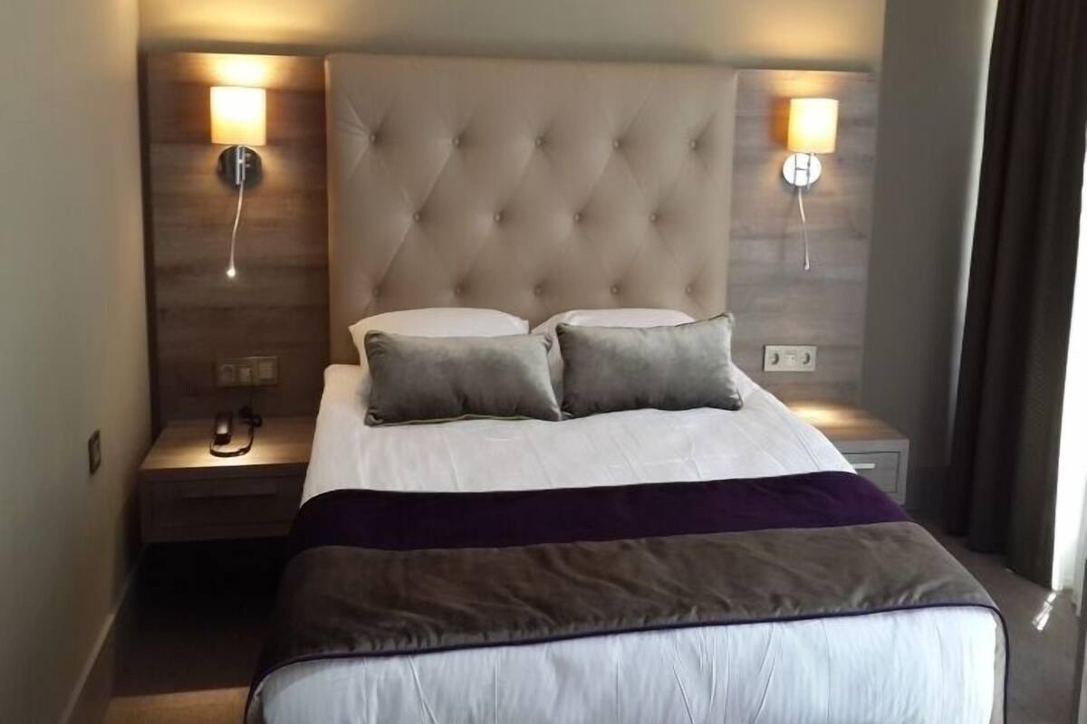 A hotel room with a tufted headboard, two bedside lamps, a bed with white linens and a purple throw, and a nightstand with a phone and a lamp.