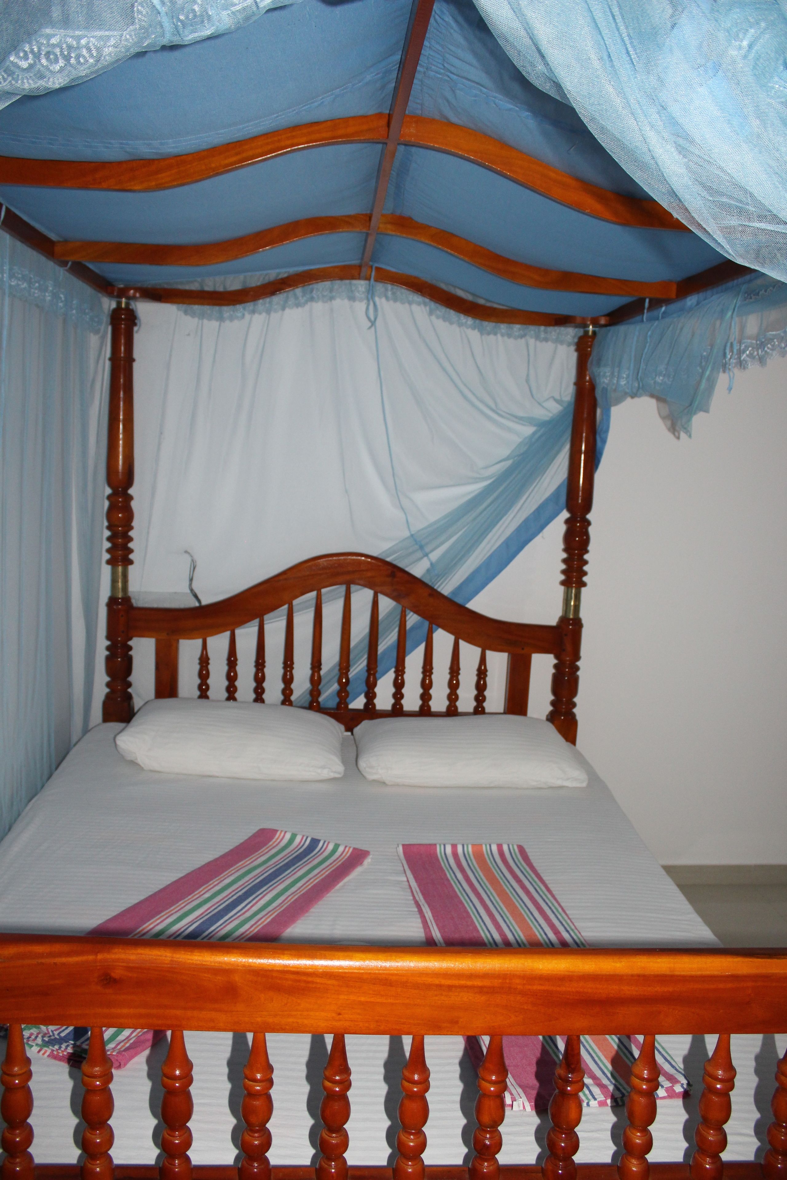 A wooden four-poster bed with a white mattress and two pillows, draped with a blue canopy.