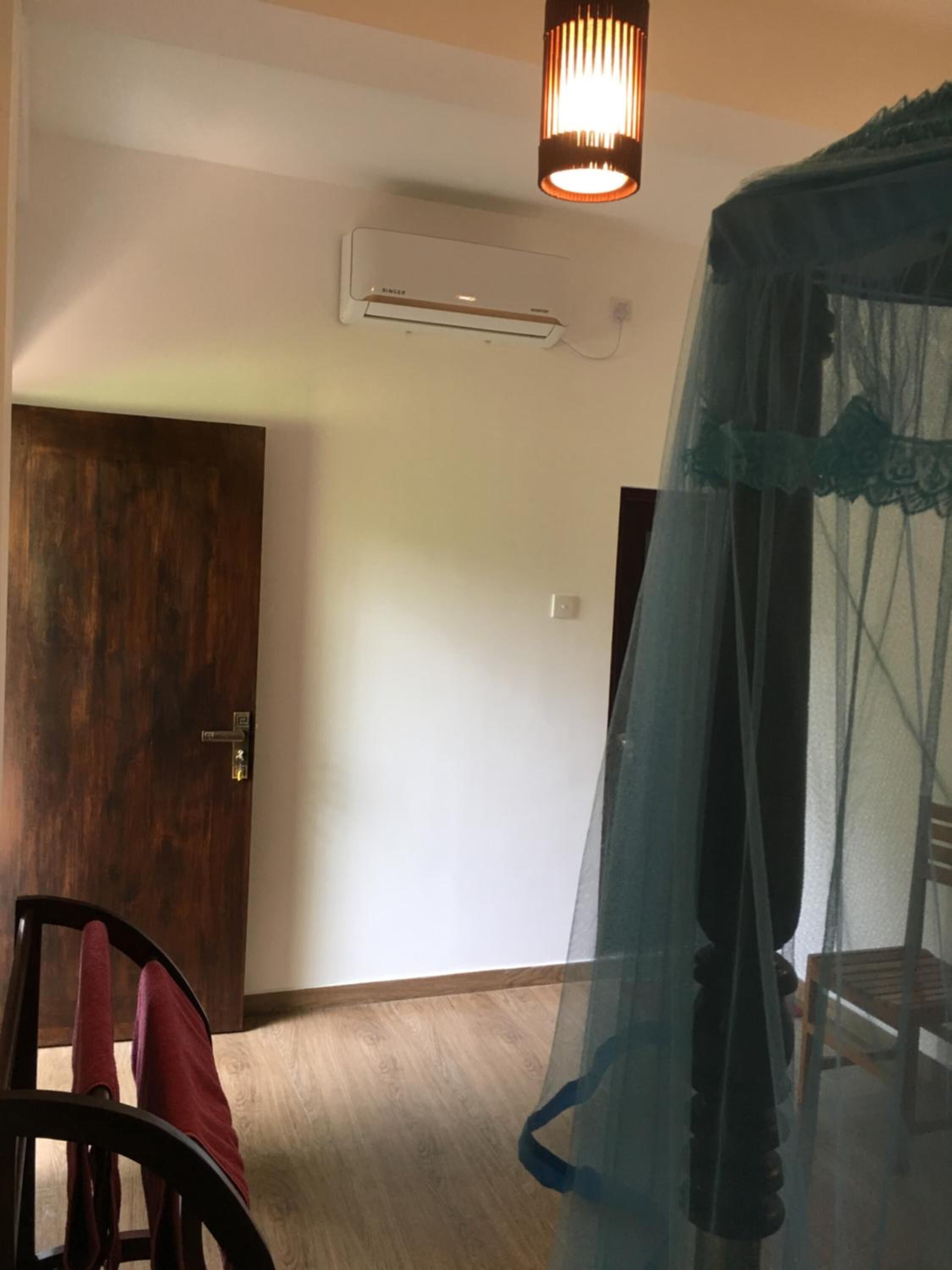 A room with a wooden door, a chair with a red cushion, a bed with a blue canopy, and an air conditioning unit on the wall.