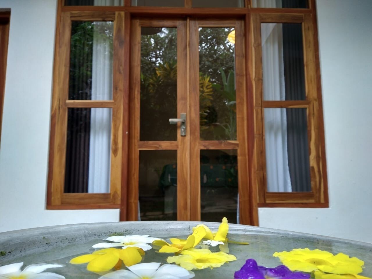 Una habitació amb un finestra gran amb marc de fusta, un sòl de formigó i pètals de flors dispersos.