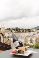 Mesa com garrafa de vinho, duas taças e prato com comida, com vista para um horizonte urbano com edifícios e um castelo.