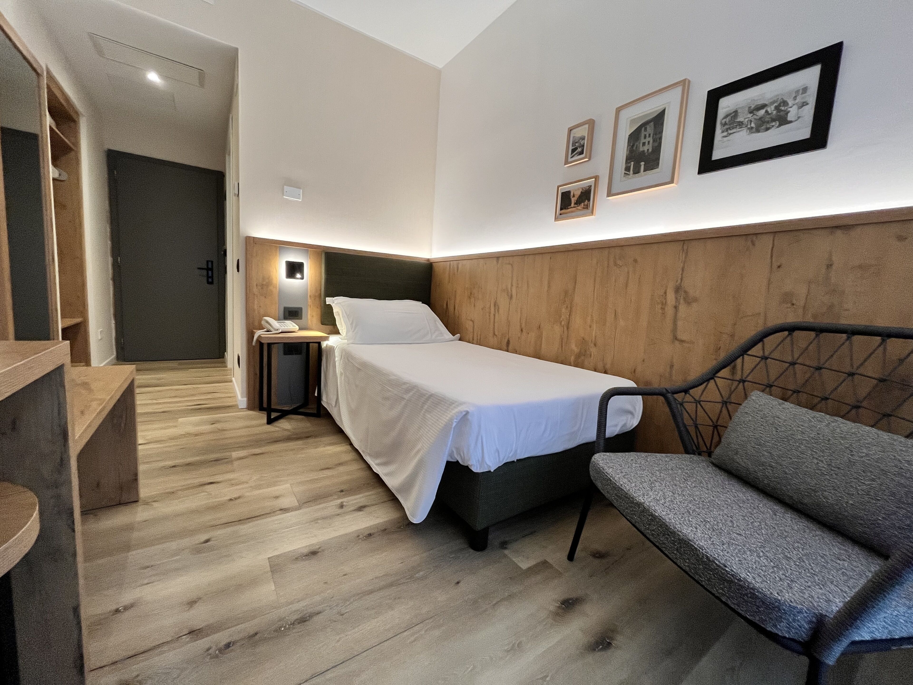 A hotel room with a bed, a chair, a nightstand, and a wall-mounted light. There are three framed pictures on the wall.