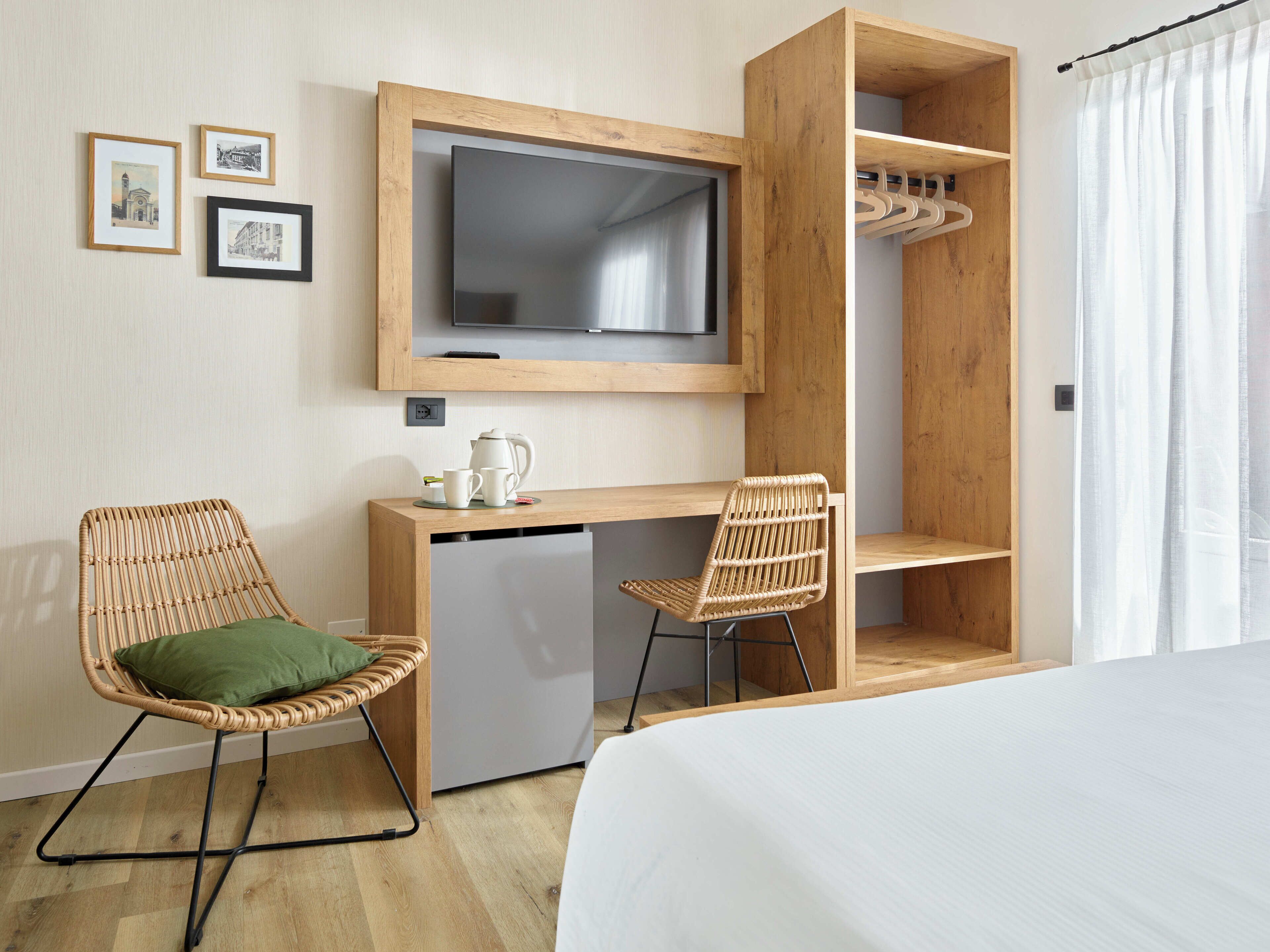 Una habitación de hotel con una cama, un escritorio con televisor, dos sillones de mimbre, una kitchenette pequeña y un armario de madera.