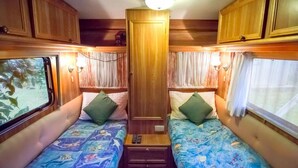 Interior compacto de uma RV com duas camas, cada uma com colcha florida e travesseiro verde. Há armários de madeira acima das camas e uma porta central.