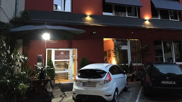 Un edificio con fachada roja, un coche blanco estacionado al frente y un letrero que dice 'Pokoje Integro'.