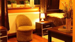 Una habitación acogedora con suelo de madera, un otomán pequeño y redondo, una mesita de noche con lámpara, una cama con cortinas oscuras y un televisor montado en la pared.