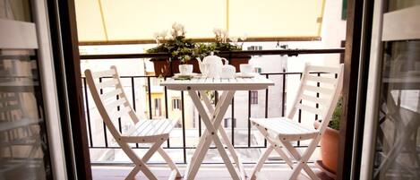 A balcony with a white table and two chairs, a potted plant, and a window with a view of buildings.