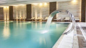 Una piscina interior moderna con fuente de agua cristalina, sillones de descanso y paredes de mármol.