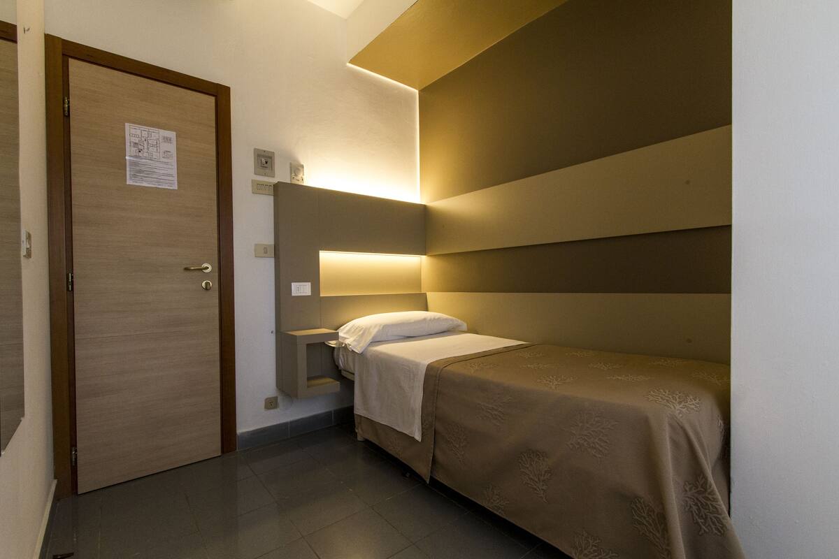 Une petite chambre à coucher, bien rangée, avec un lit simple, une table de chevet et une porte sur laquelle est affiché un avis.
