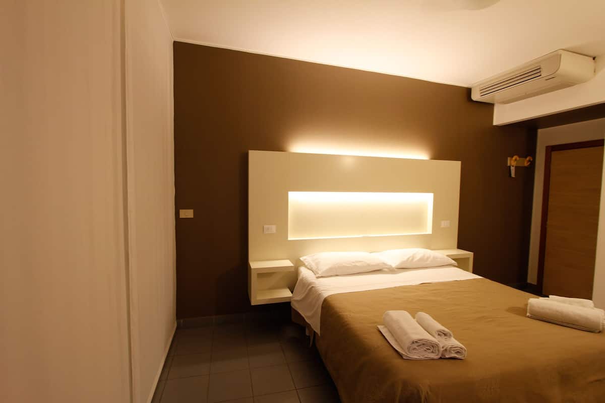 Une chambre d’hôtel avec un grand lit, une table de chevet, un climatiseur fixé au mur et une porte avec une poignée.