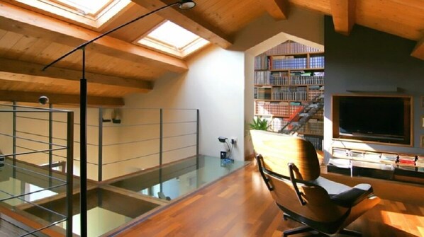 A spacious loft with a high ceiling, skylights, a glass railing, a wooden floor, a comfortable chair, a bookshelf, and a television.