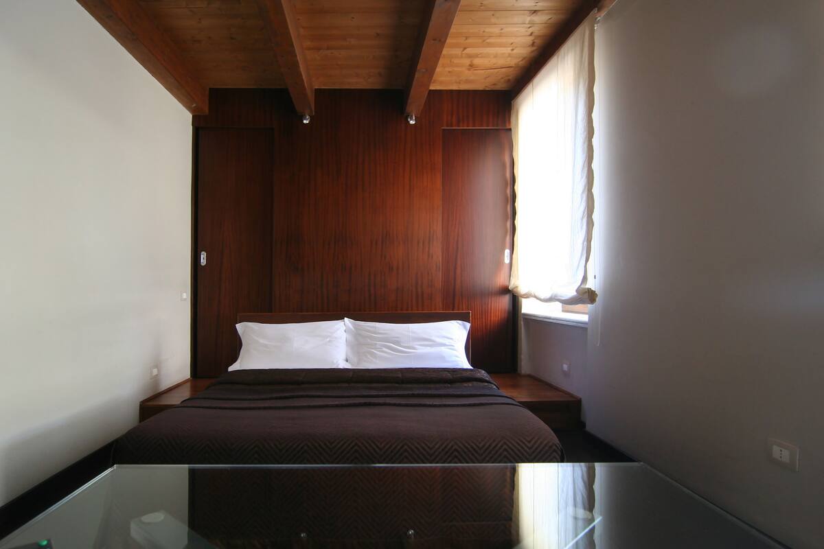 A bedroom with a wooden headboard, a glass table, and a window with curtains.