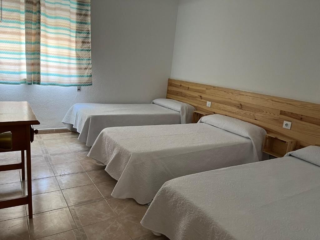 Une chambre avec trois lits simples, une tête de lit en bois, une petite table et une fenêtre avec des rideaux.