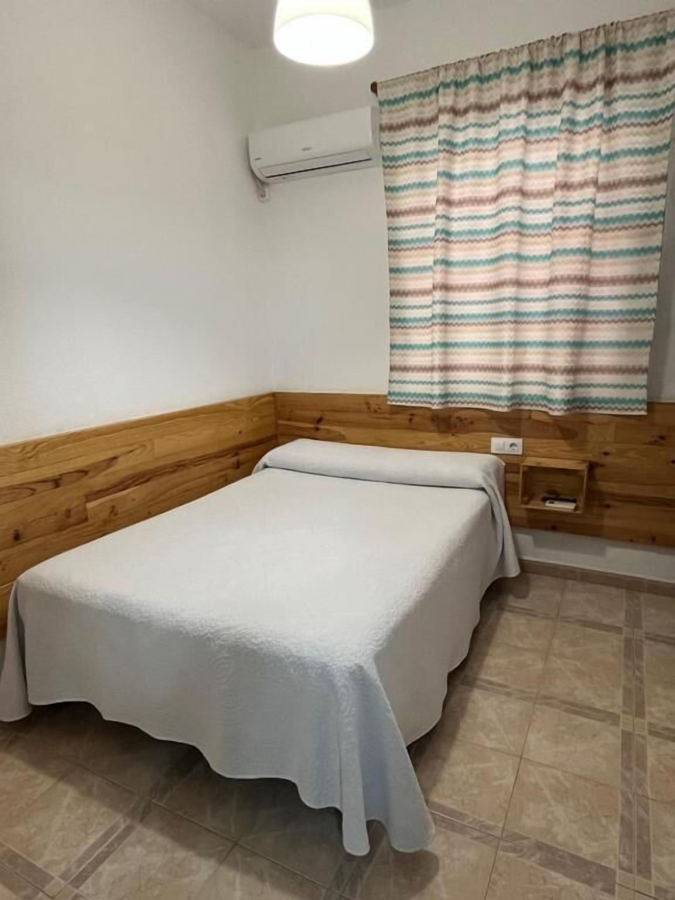 Una cama individual en una habitación con paneles de madera, una mesita de noche pequeña y una ventana con cortinas a rayas.