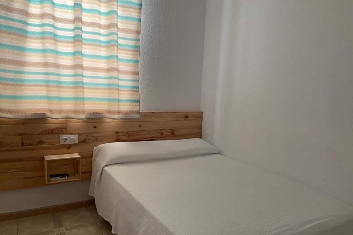 A small, single-bed room with a wooden headboard, a bedside table, and a window with striped curtains.