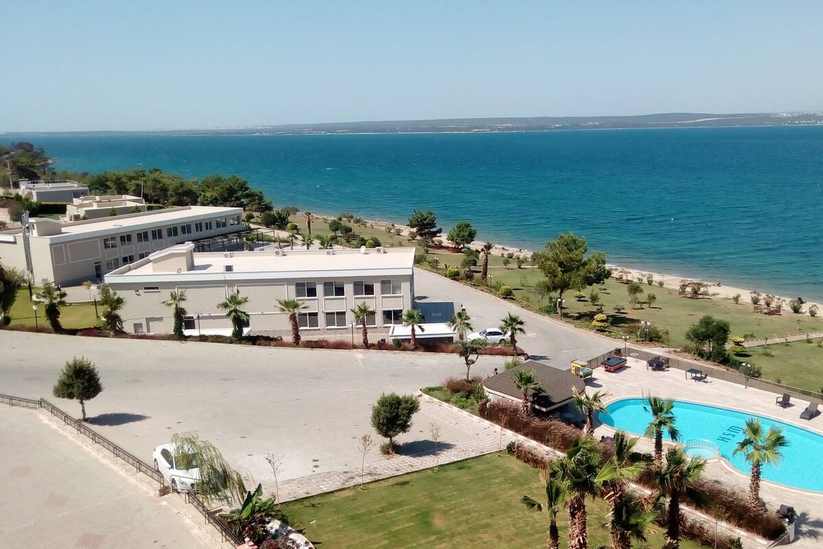 Um edifício moderno com piscina, rodeado por um jardim paisagístico com palmeiras e vista para o mar.