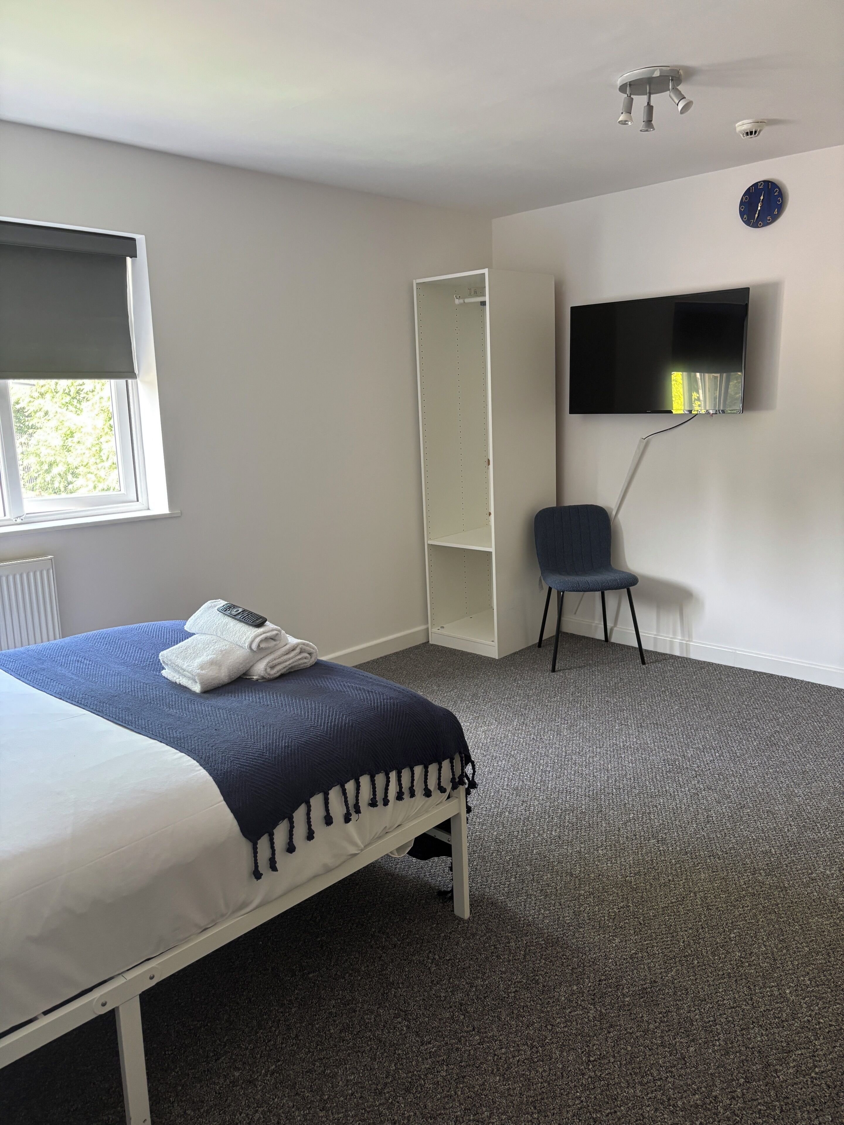 Ein Hotelzimmer mit einem Bett, einem an der Wand befestigten Fernseher, einem Sessel, einem Kleiderschrank und einem Fenster.
