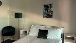 A neatly made bed with white linens, a dark green chair, a floor lamp, and a painting on the wall. - Tlk Apartments And Hotel (Orpington)