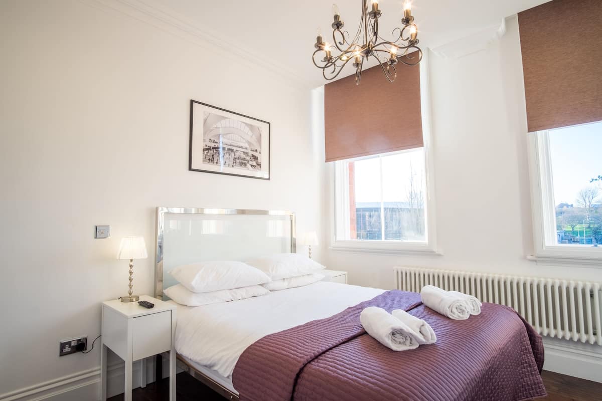 A hotel room with a bed, a nightstand, a lamp, a framed picture on the wall, and a window with a view.