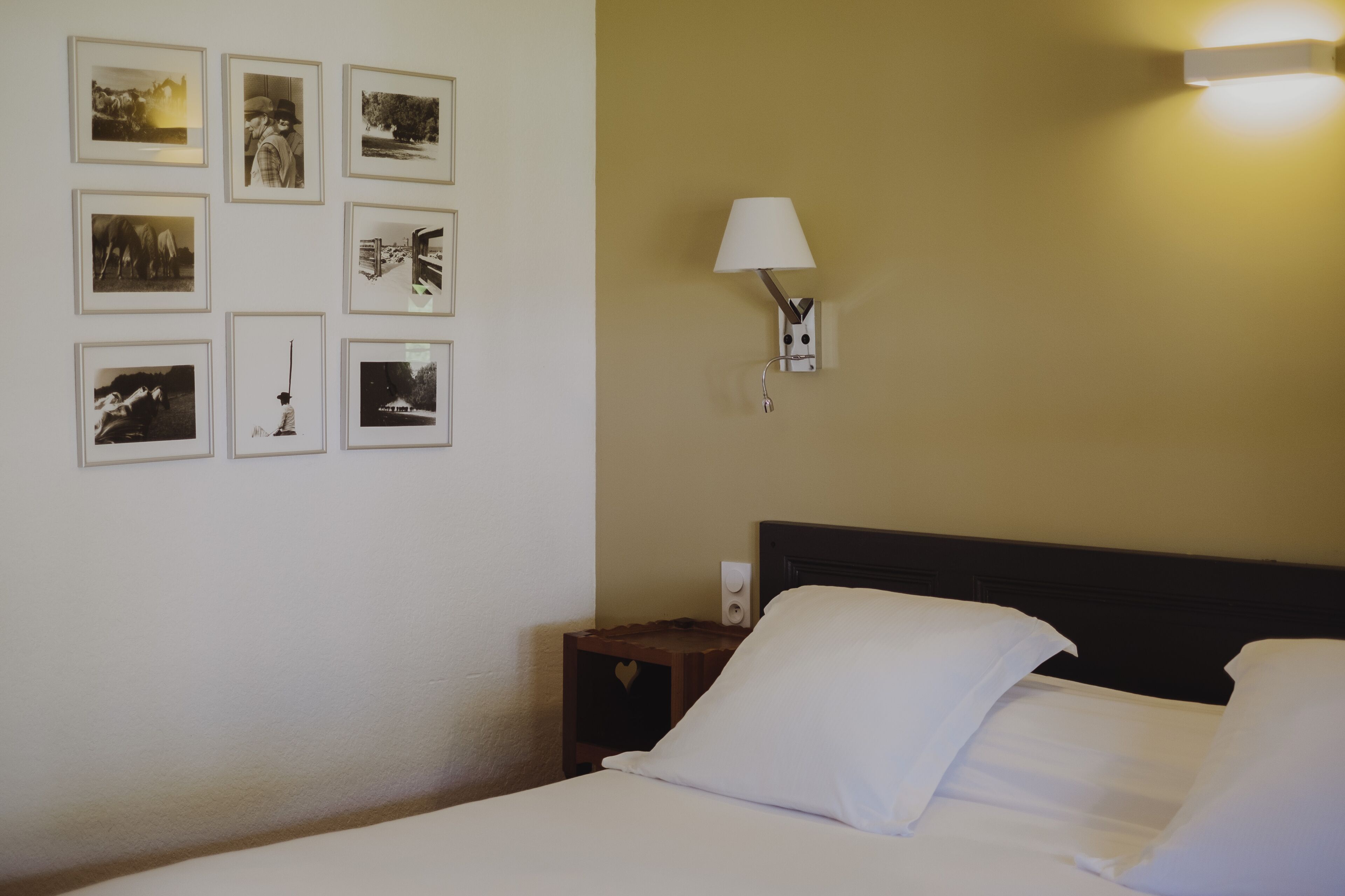 Una habitació d'hotel amb un llit ben fet, una tauleta de nit amb una làmpada, i una paret decorada amb fotografies emmarcades.