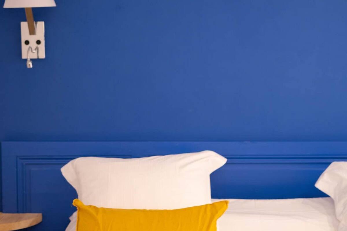 Een bed met witte en gele kussens, een donkergroene deken en een blauwe muur met een witte lamp.