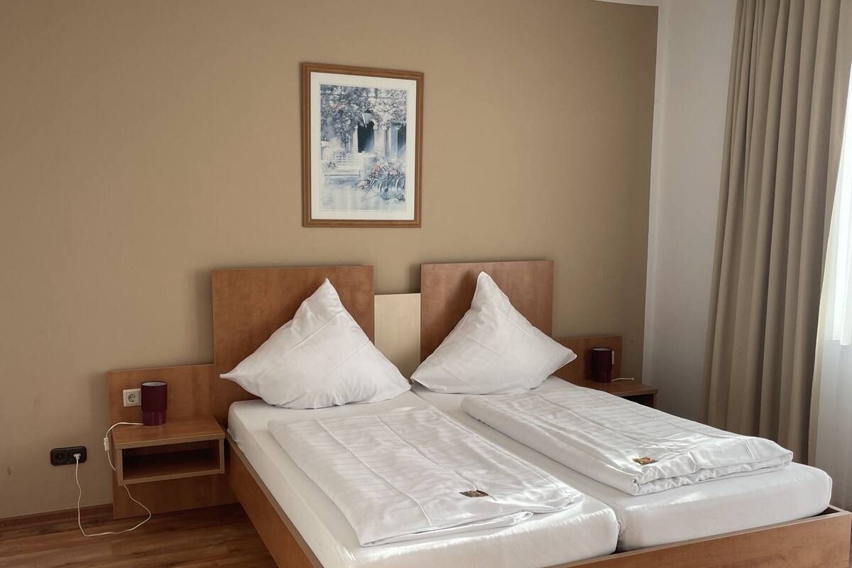 Una cama doble con cabecera y piecera de madera, dos almohadas blancas, una mesita de noche con una taza roja y un cuadro en la pared.