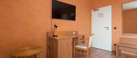 Une pièce comprenant un bureau en bois, une chaise, une petite table en osier, un téléviseur fixé au mur et une porte sur laquelle est accroché un tableau d’affichage.
