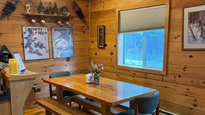 Puinen mökki, jonka sisustuksessa on ruokapöytä, penkit, ikkuna, jossa on sälekaihtimet, kehyksiin laitettuja urheilulehtiä ja pieni hylly koriste-esineitä varten.